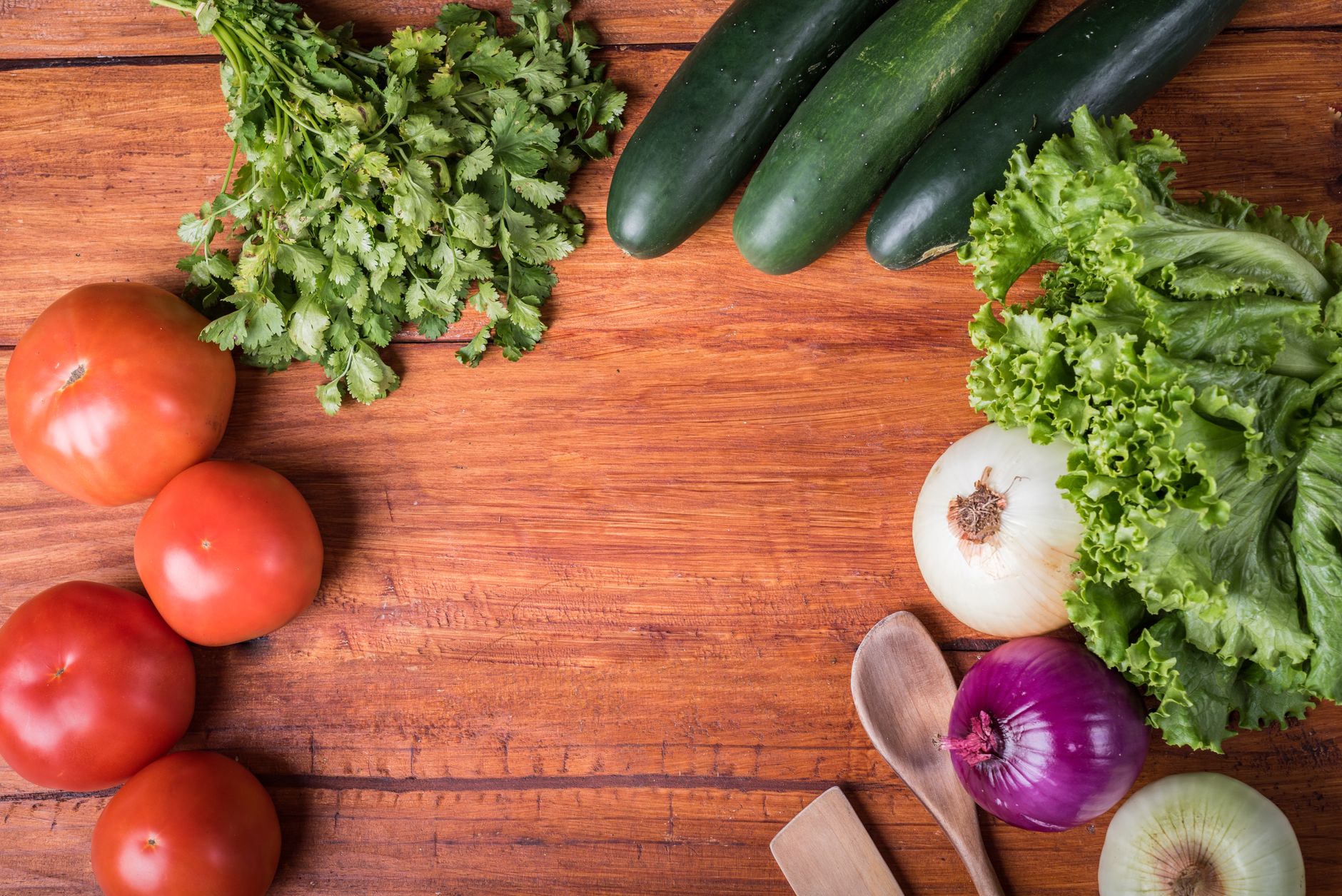 Top view of fresh vegetables including tomatoes, cilantro, lettuce, cucumbers, and onions on a wooden surface.